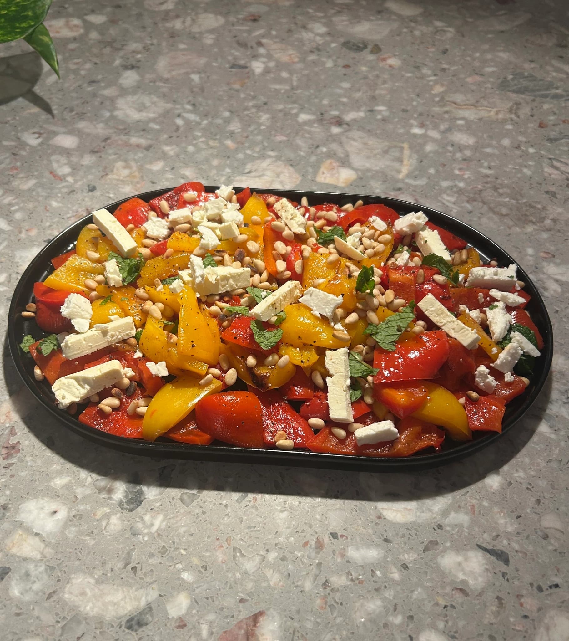 Grilled bell pepper salad in warm tones with red, yellow, and orange peppers, topped with crumbled feta cheese, toasted pine nuts, and fresh parsley. Served on a rustic ceramic platter with olive oil sheen, Mediterranean summer light